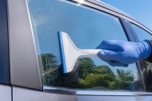 Main gantée utilisant une raclette en plastique pour nettoyer la résine sur une vitre de voiture.